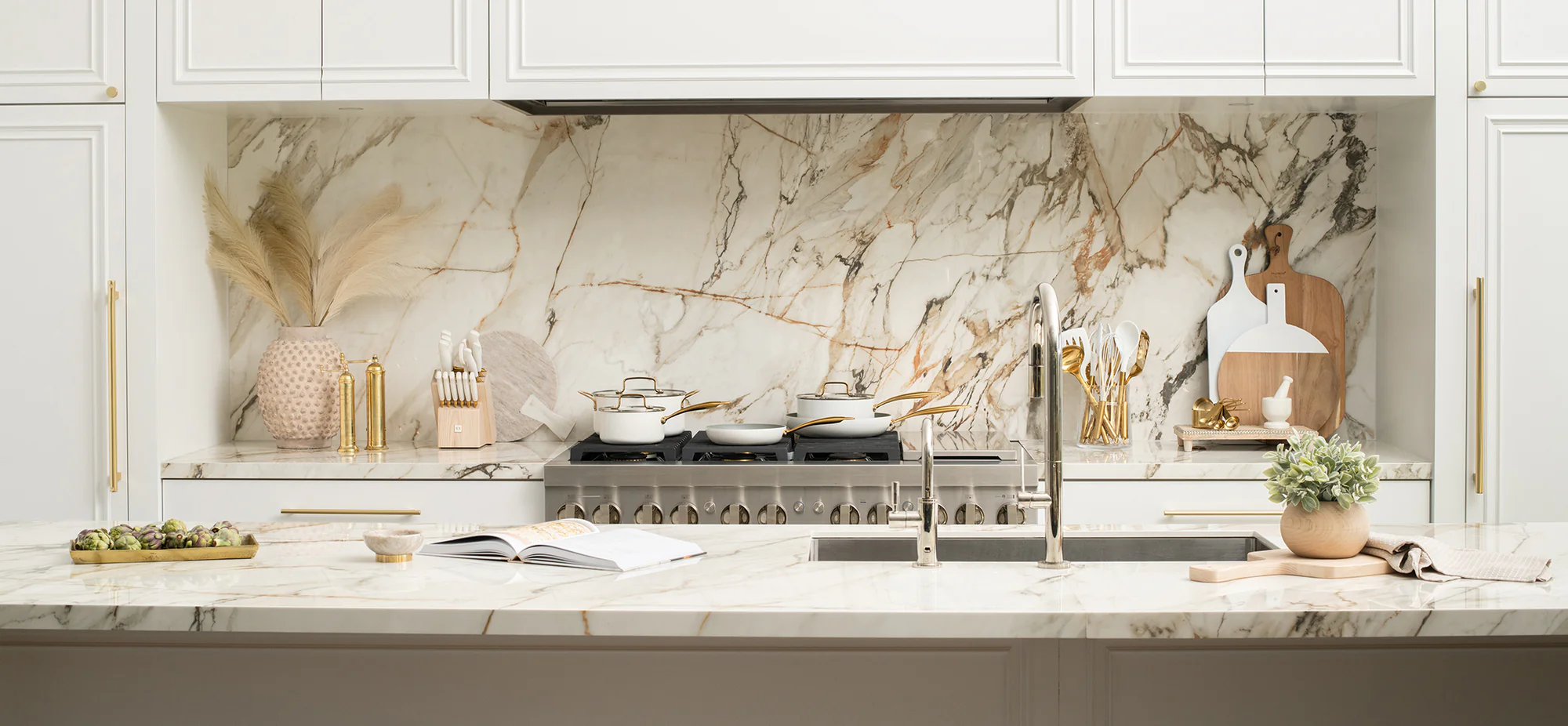Wide shot of a modern kitchen featuring Spring Chef cream cookware with gold handles, knife block, utensils, and open cookbook on a marble island.